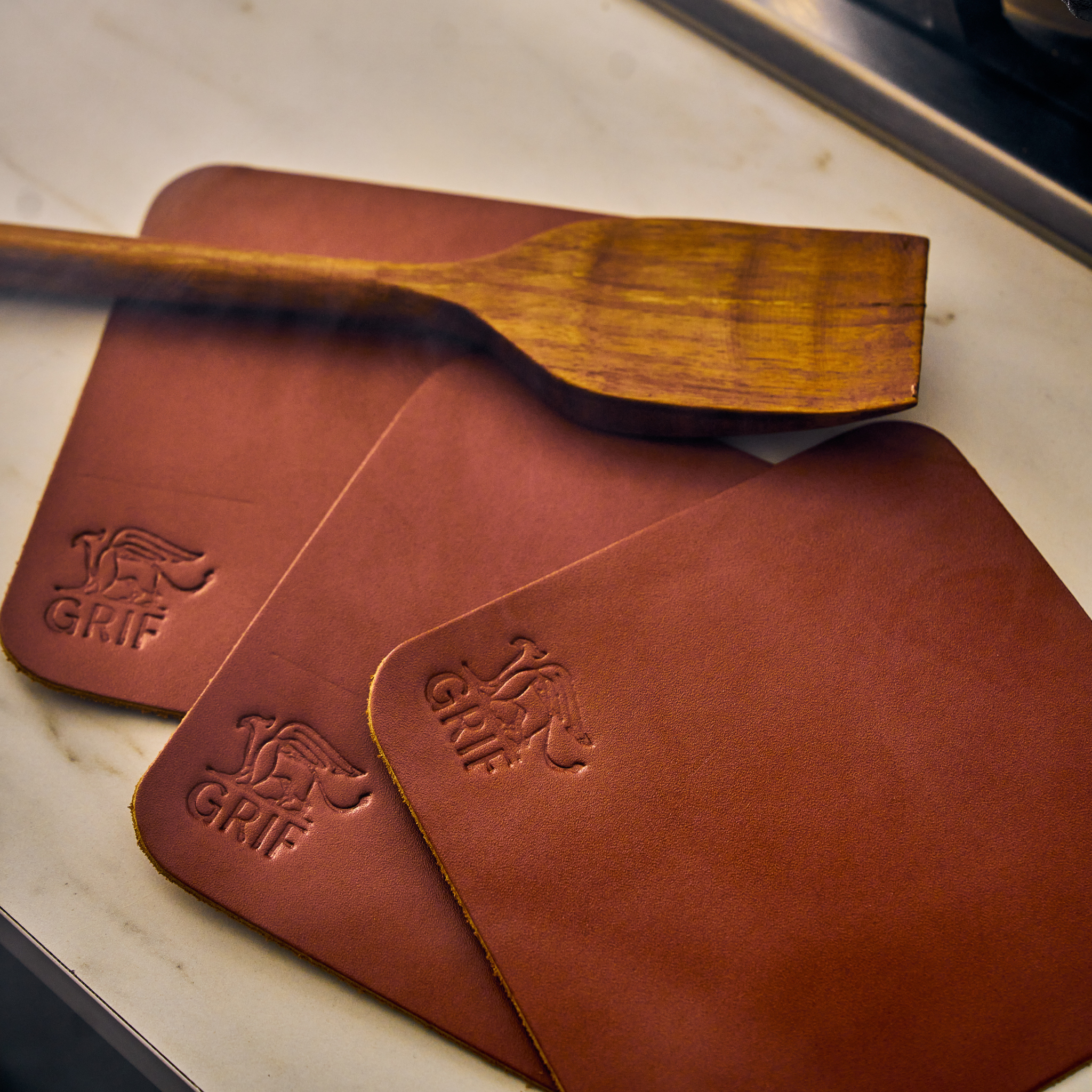 A Dutch oven and grill leather handle/coaster set by GRIF, featuring a brown leather handle cover and a matching coaster.