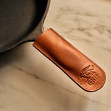 Close-up of a cast iron skillet with a brown genuine leather handle cover by GRIF, featuring an embossed winged creature logo and the text "GRIFF."